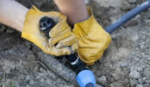 Gloved hands performing irrigation system repair on a broken pipe using a connector in a dirt field.