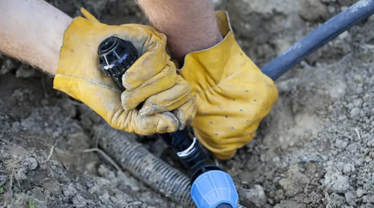 Gloved hands performing irrigation system repair on a broken pipe using a connector in a dirt field.